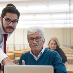 An elderly man learning computer skills with a teacher in a classroom setting, showcasing education through technology.