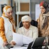 Three professional women in hijabs discussing documents at their office desk. Teamwork and collaboration concept.