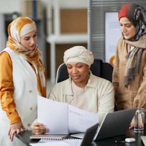 Three professional women in hijabs discussing documents at their office desk. Teamwork and collaboration concept.