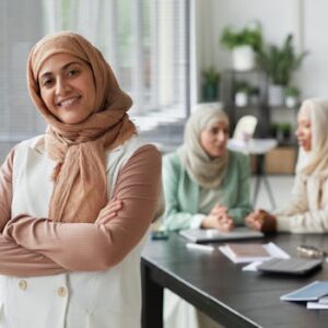 Smiling Muslim woman in hijab standing confidently in an office with colleagues in the background.
