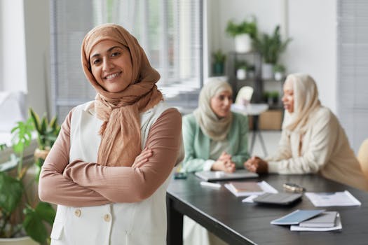 Smiling Muslim woman in hijab standing confidently in an office with colleagues in the background.