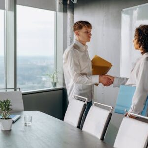 Two colleagues shaking hands in a bright conference room with documents.