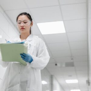 A focused female scientist in a lab coat writes notes in a laboratory hallway, showcasing professionalism in research settings.