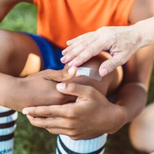 A close-up view of an adult applying a band aid to a child's wounded knee outdoors during the day.