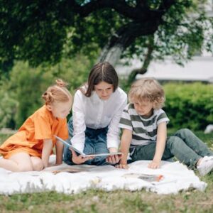 Three kids reading a book on a picnic blanket in a park under a tree.