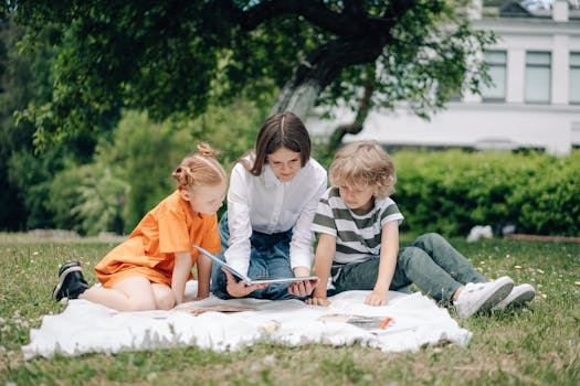 Three kids reading a book on a picnic blanket in a park under a tree.