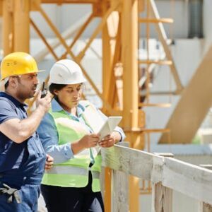 Two construction workers in hardhats discuss plans using a tablet and walkie talkie on-site.