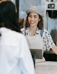 Smiling female barista in apron and hat serves customer in cafe.