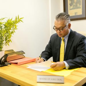 An adult man in business attire working at a desk with documents in an office environment.