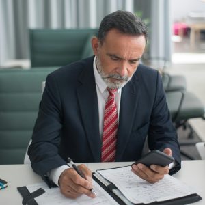 A professional businessman in a suit reads documents and checks his phone at a desk.