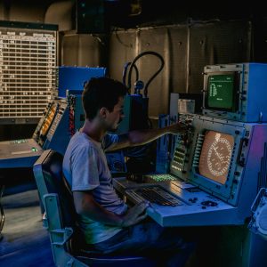 A man working at control panels in a dimly lit industrial room with various monitors.