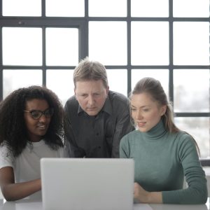 Positive focused multiracial colleagues read information from laptop while teamwork on project in office with industrial interior against big window at daytime