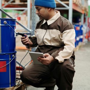 Man in beanie inspects warehouse drum, using a tablet for inventory.
