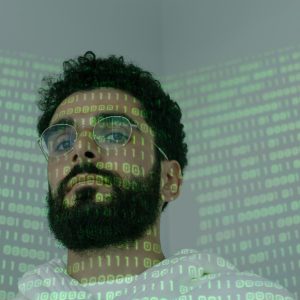 A man with a beard and eyeglasses looking at the camera, illuminated by binary code projection.