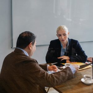 Smart colleagues in formal outfit discussing business while sitting at table with cups of coffee in office with whiteboard