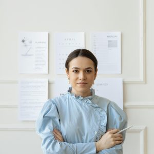 Professional woman in stylish attire standing in a modern office setting with crossed arms.