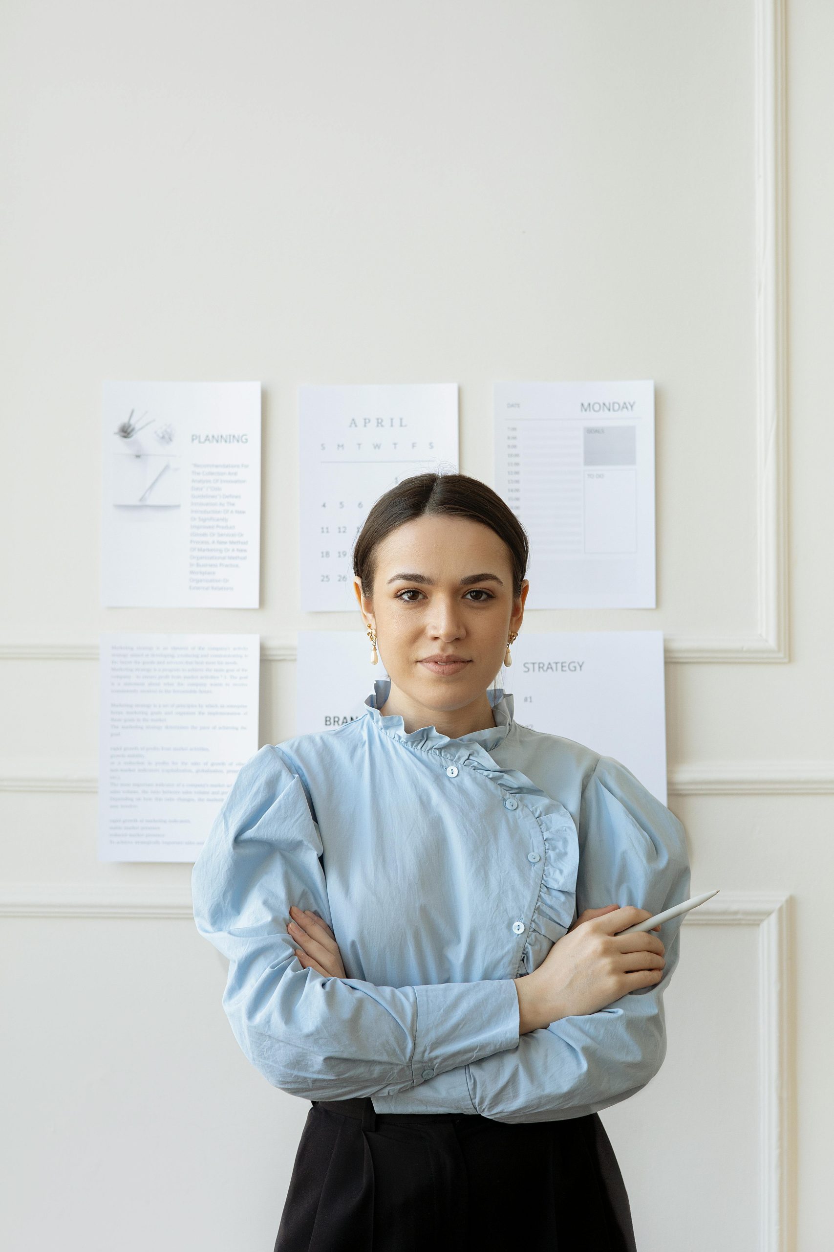 Professional woman in stylish attire standing in a modern office setting with crossed arms.