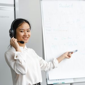 Smiling woman in a white shirt presenting with a headset and flipchart in an office environment.
