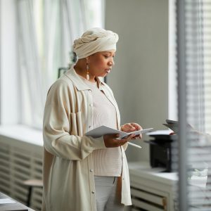 Businesswoman in headwrap reviewing documents near office window. Professional workspace setting.