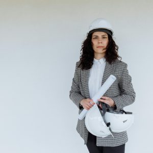 Engineer in formal attire holding blueprints and hard hats, standing against a white wall.
